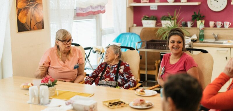 Elderly residents and caregivers enjoying a meal and conversation in a cheerful dining space.